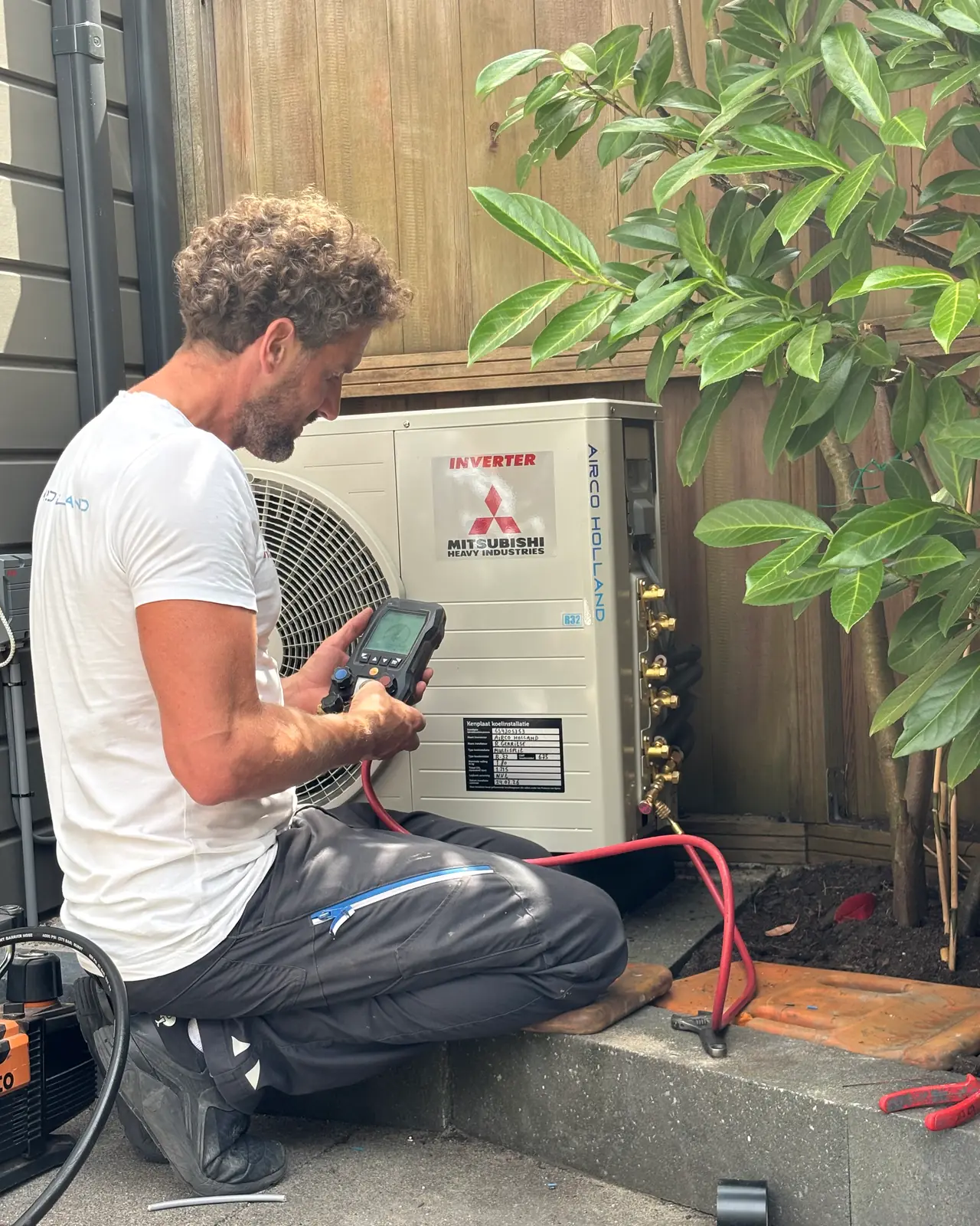 Rein zit bij een buitendeel in een tuin en leest de meter af die met een rode slang is aangesloten.